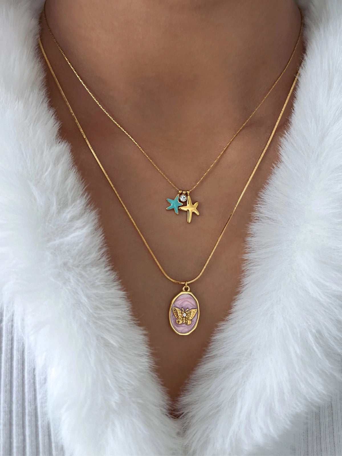 Flora Butterfly Necklace – On Model Styled Shot-Two gold necklaces with star and butterfly pendants on a white fur background
