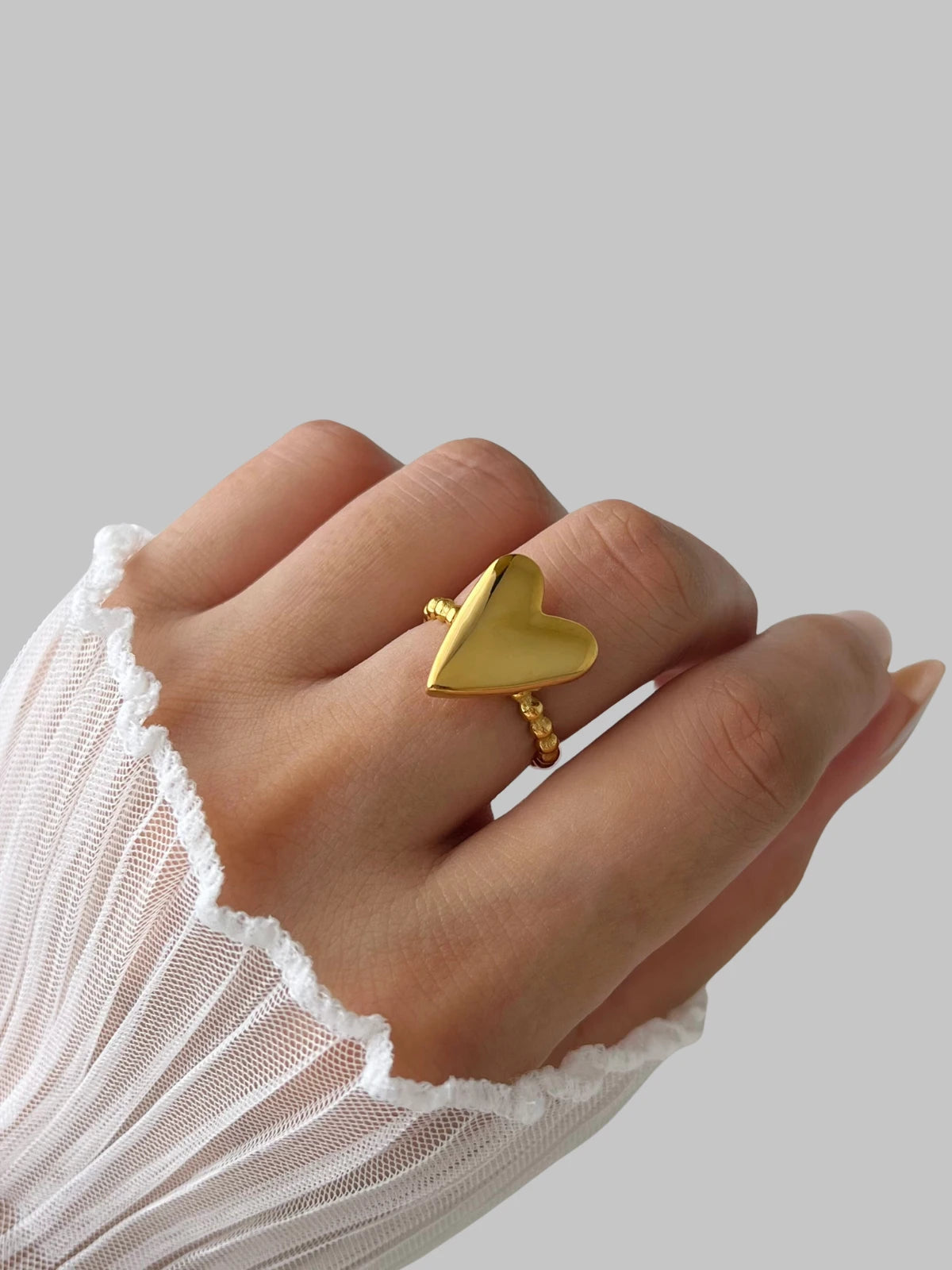 Love Vibe Ring – On Model Styled Shot-Gold heart-shaped ring on a hand with a white lace sleeve against a light gray background