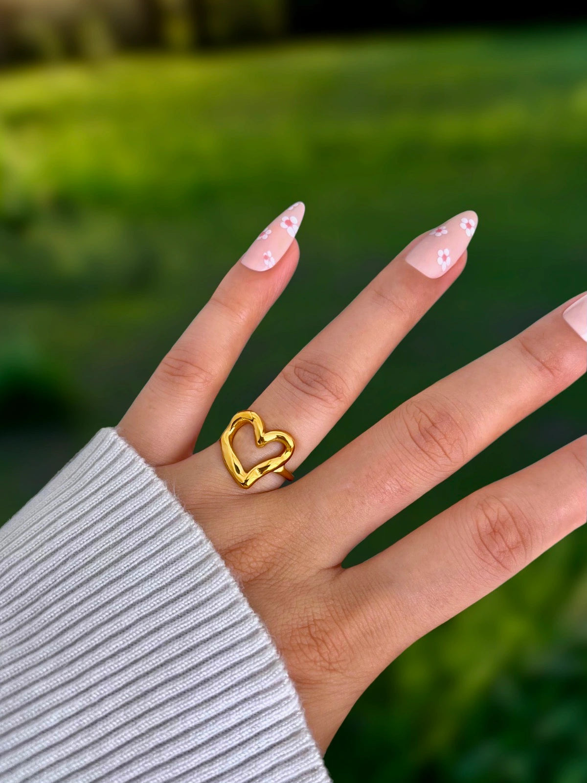 My Heart Loop Ring – Worn on Finger-Hand wearing a gold heart-shaped ring with a blurred green background