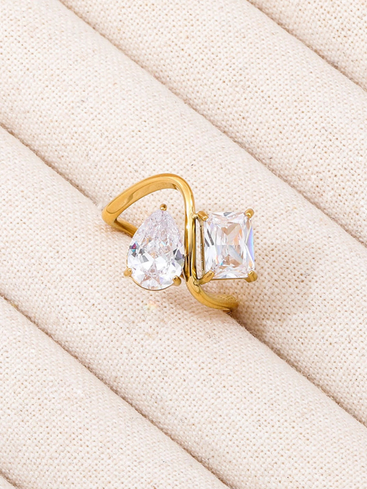 Water_Drop_Ring_Front_View_1. Gold ring with two large clear gemstones on a beige fabric background