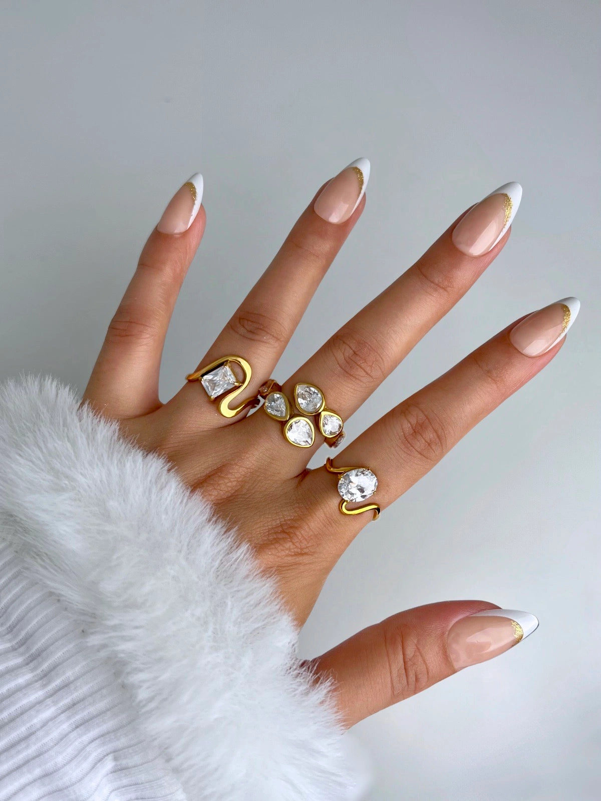 noa-ring-closeup-detail-shine-with-me-Hand wearing multiple gold rings with clear gemstones on a light gray background