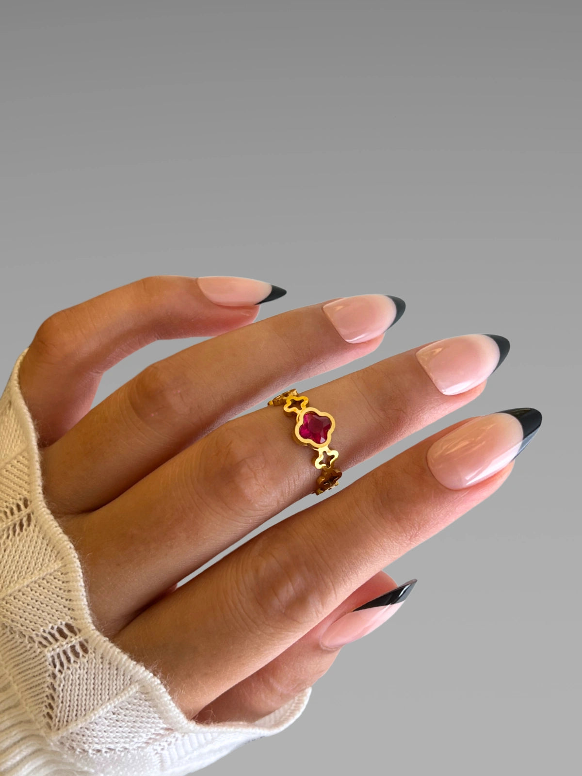 Amara Clover Ring | Shine With Me Hand wearing a gold ring with a red gemstone on a gray background