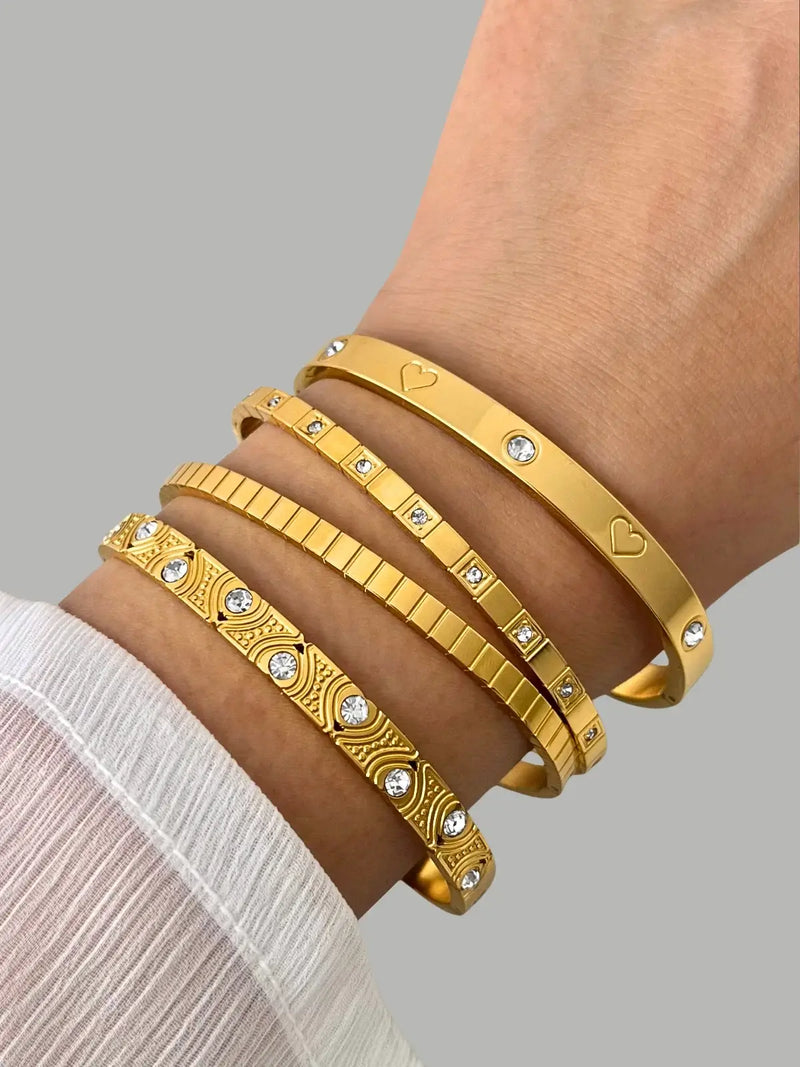 Bella Vista LOVE BANGLE worn on wrist — 18K gold-plated stainless steel bangle, width 65 mm, by Shine With Me, styled in Gold variant. Gold bracelets with heart and diamond designs on a wrist against a gray background