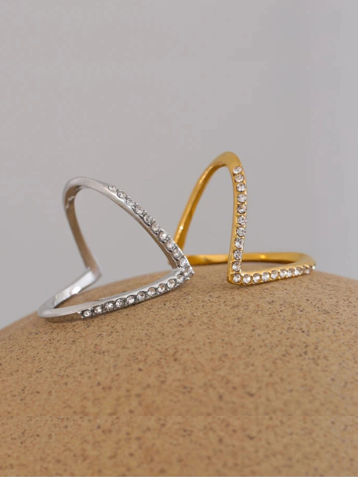 Close-up of Crush Vow Ring by Shine With Me showing gold-plated finish, stainless steel base and cubic zirconia accents.-Two gold and silver open rings with small stones on a beige surface.