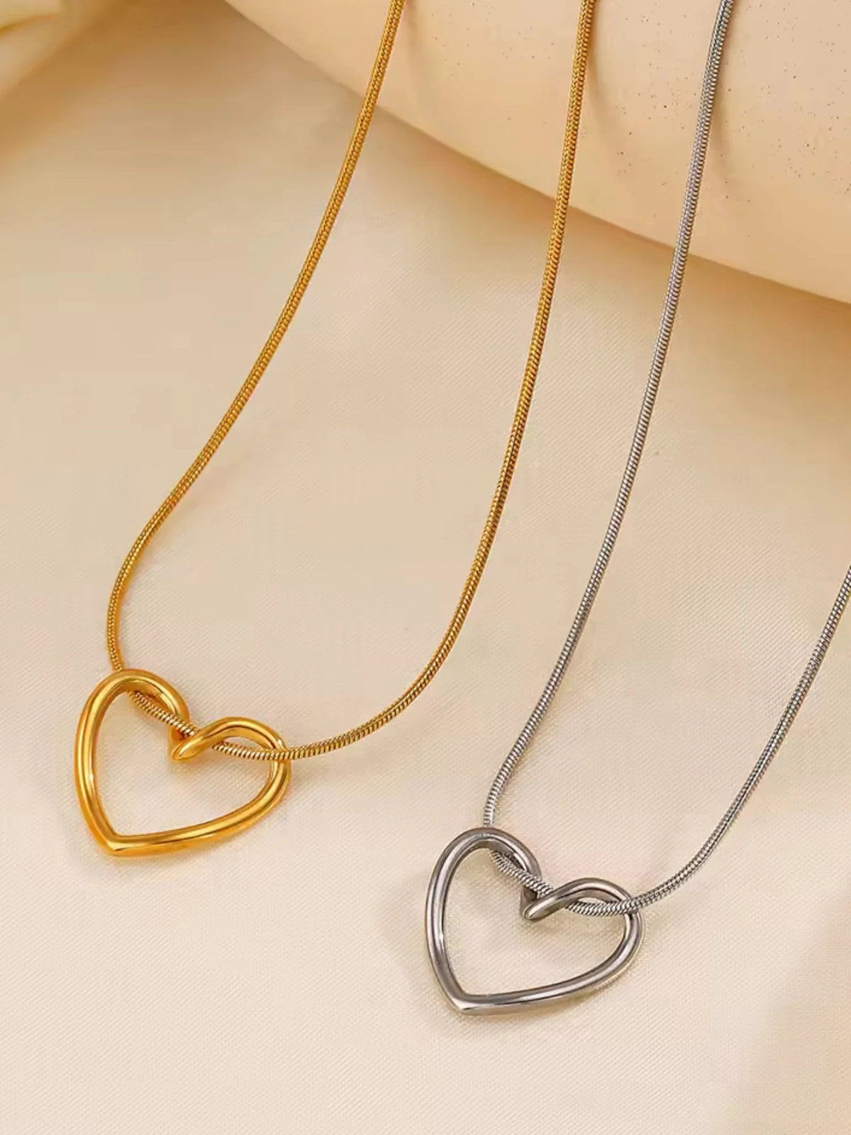 Darla Heart Necklace – Close‑Up Detail-Two heart-shaped necklaces, one gold and one silver, on a beige background.