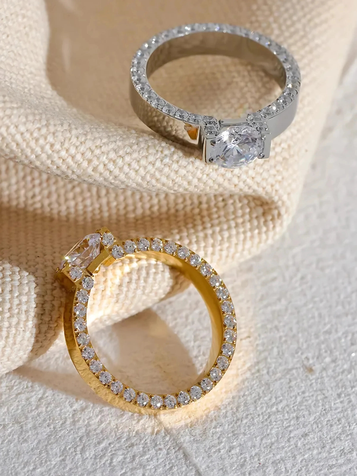 Dolores Ring – Two diamond rings, one gold and one silver, on a textured beige surface.