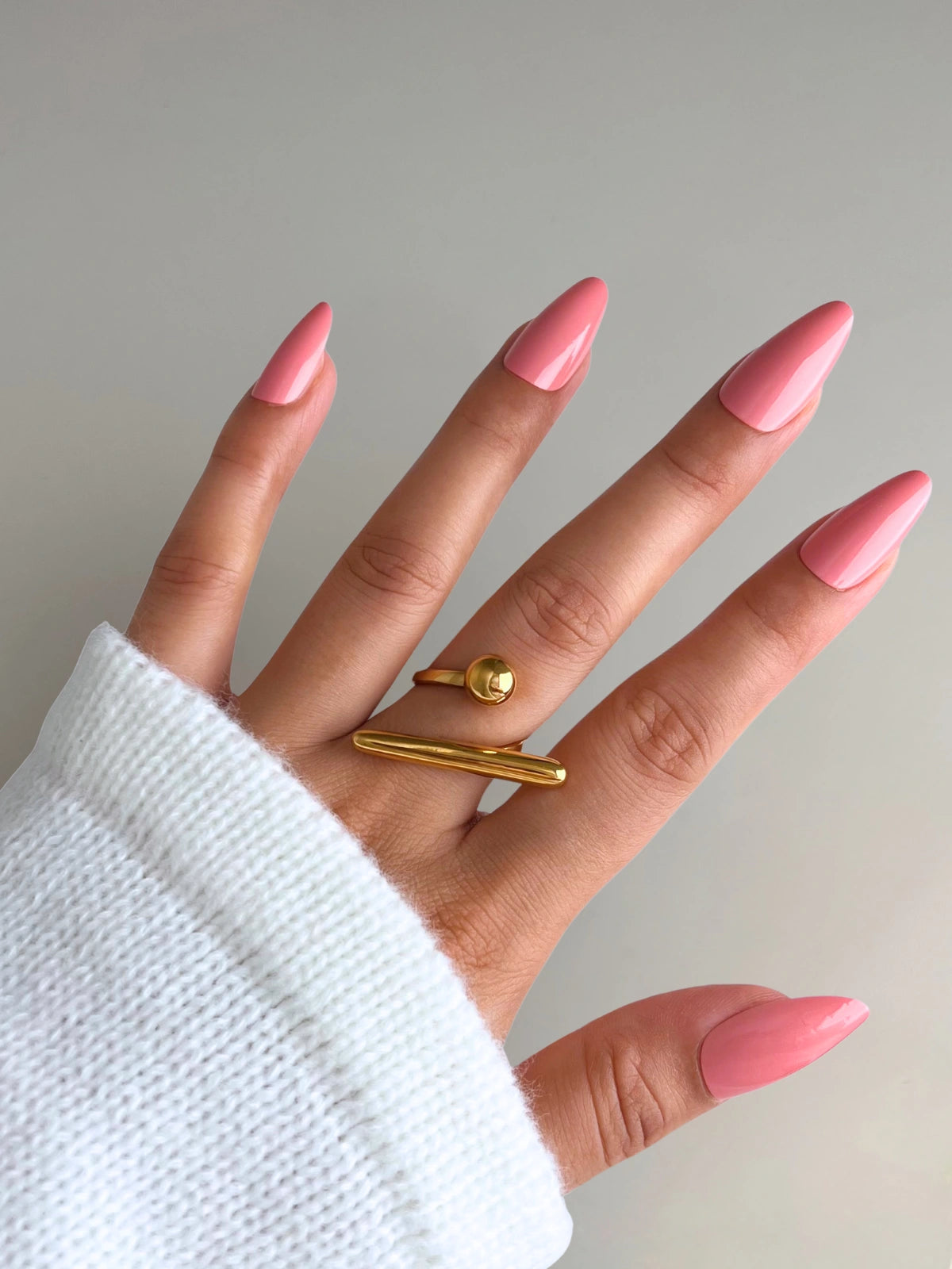 Ellowen Ring Hand with pink nails and a gold ring on a plain background