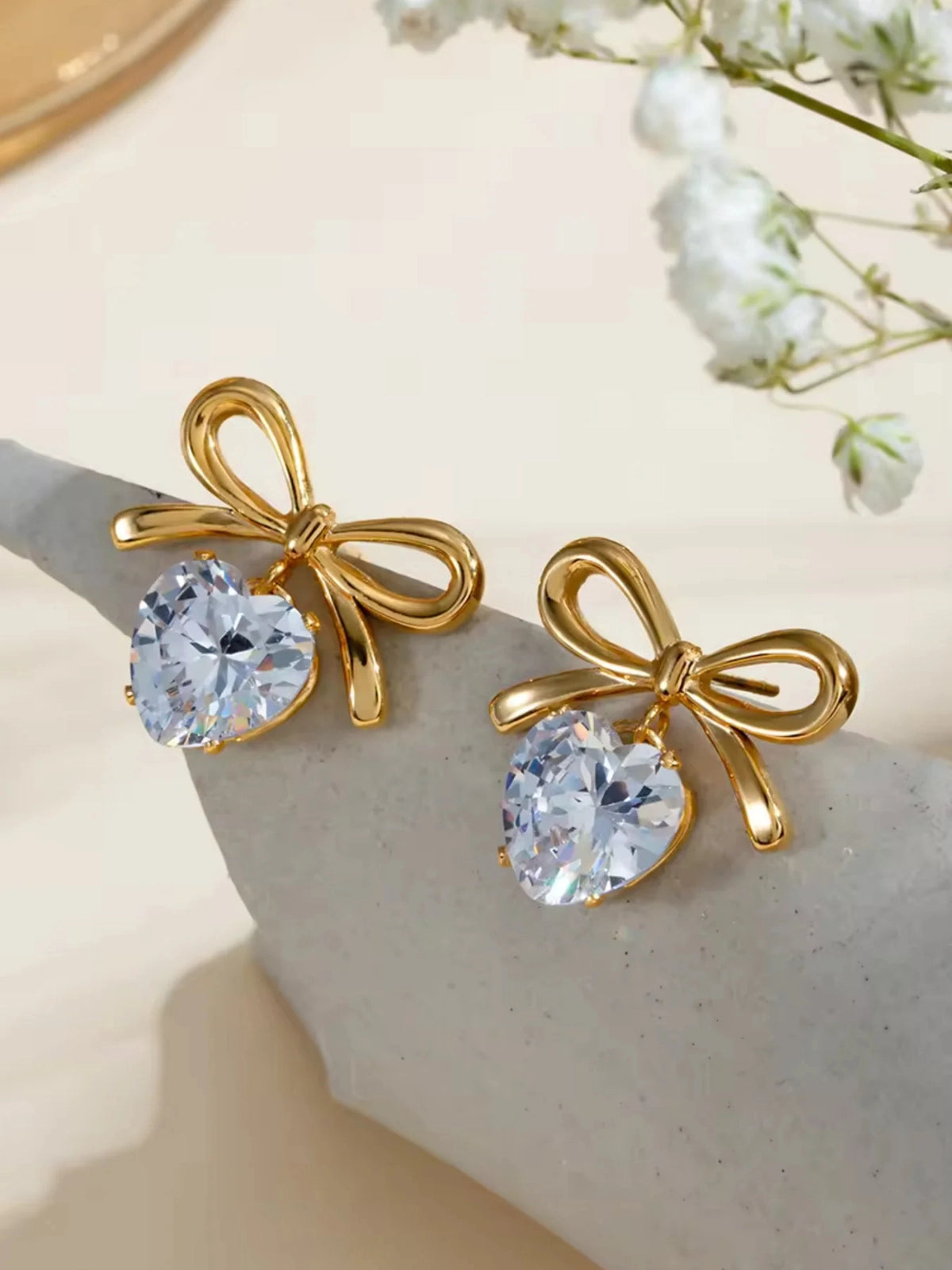Heartbow Earring – Close‑Up Detail-Gold bow-shaped earrings with clear gemstones on a light surface with flowers.