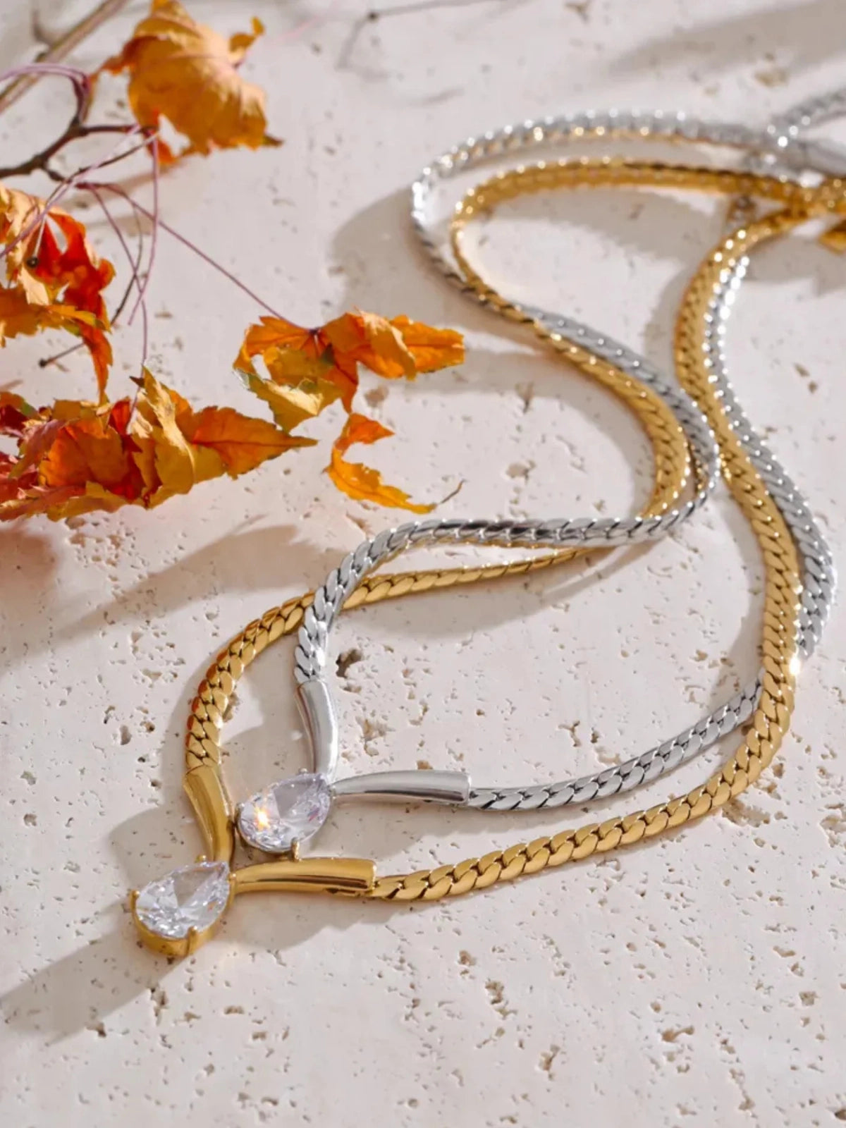 Imber Necklace – Close‑Up Detail-Gold and silver necklaces with crystal pendants on a light surface with orange leaves.