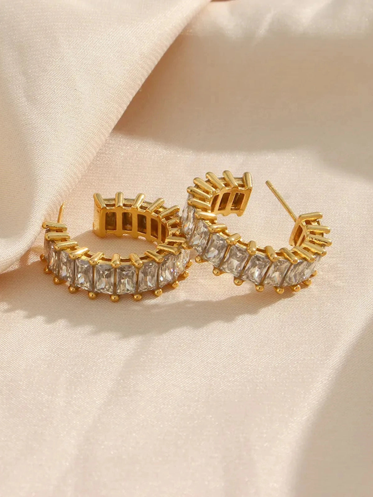 Close‑up of Ingrid Hoop Earring by Shine With Me showing gold‑plated finish, stainless steel base and cubic zirconia accents-Gold hoop earrings with clear gemstones on a beige background