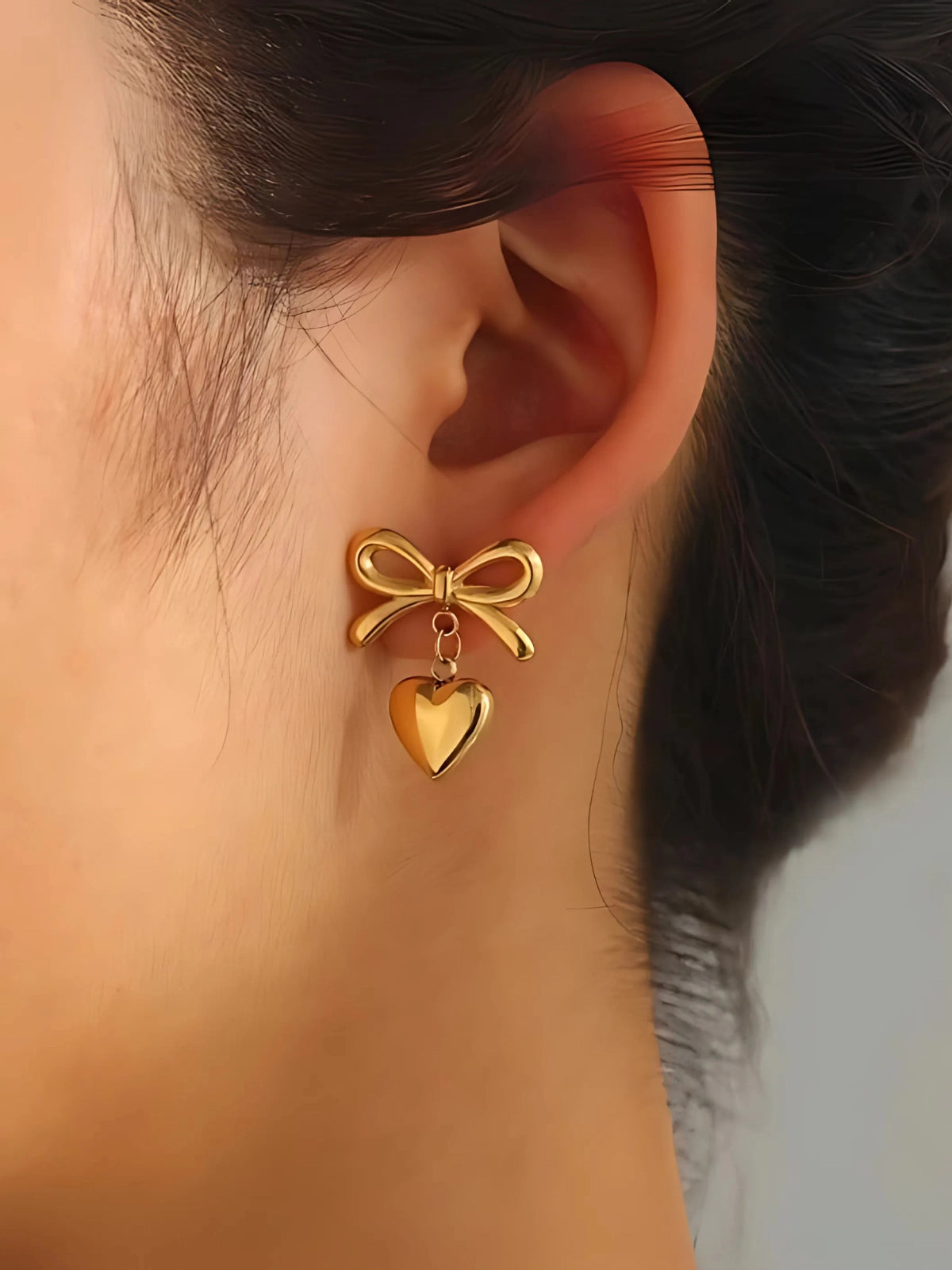 Kesha Bow Earring worn on ear — 18K gold‑plated stainless steel bow‑design by Shine With Me, styled for chic and feminine look-Gold bow and heart earring worn on an ear with a blurred background
