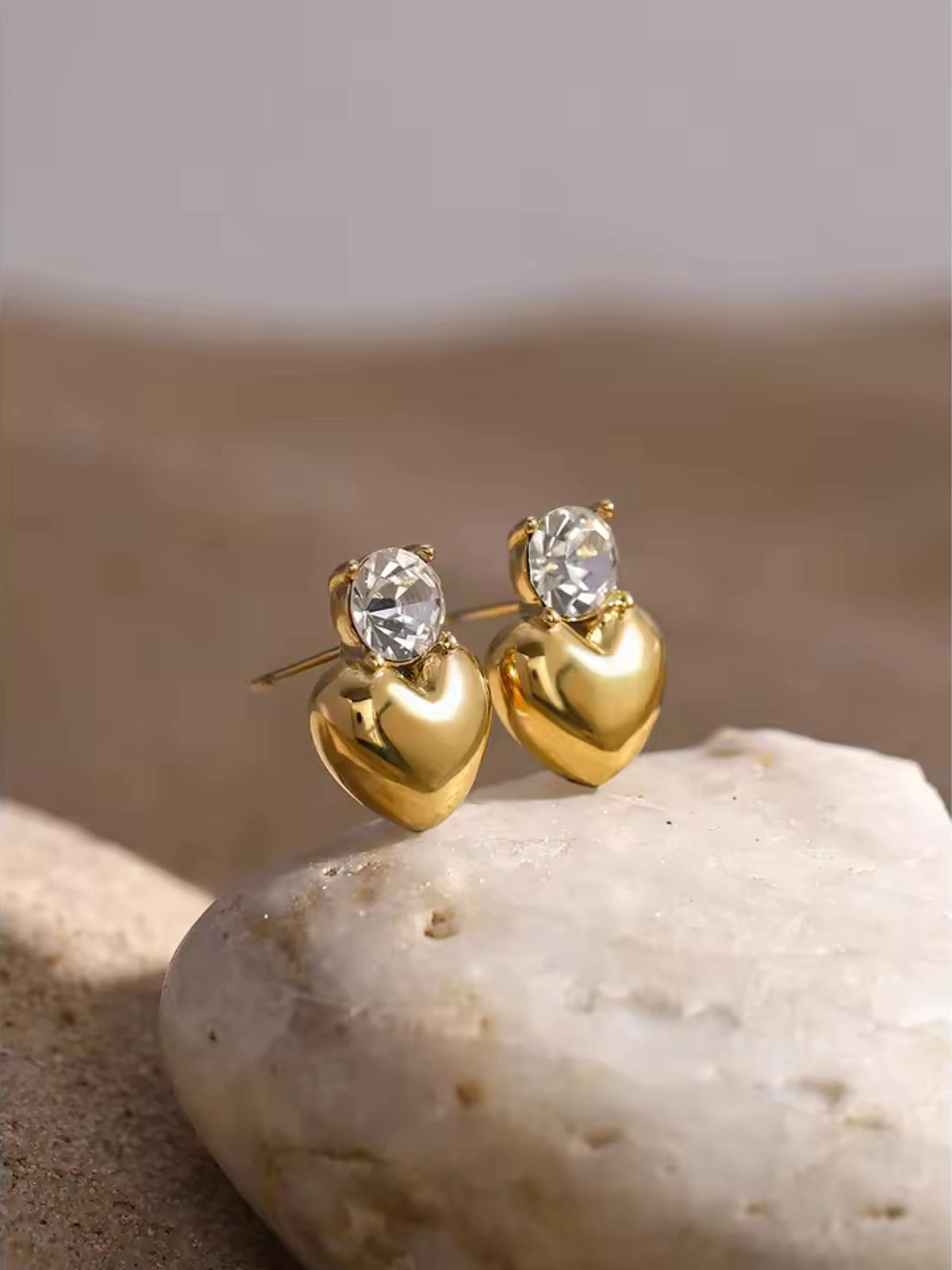 Loma Studs Earring – Close‑Up Detail-Gold heart-shaped earrings with clear gemstones on a stone surface
