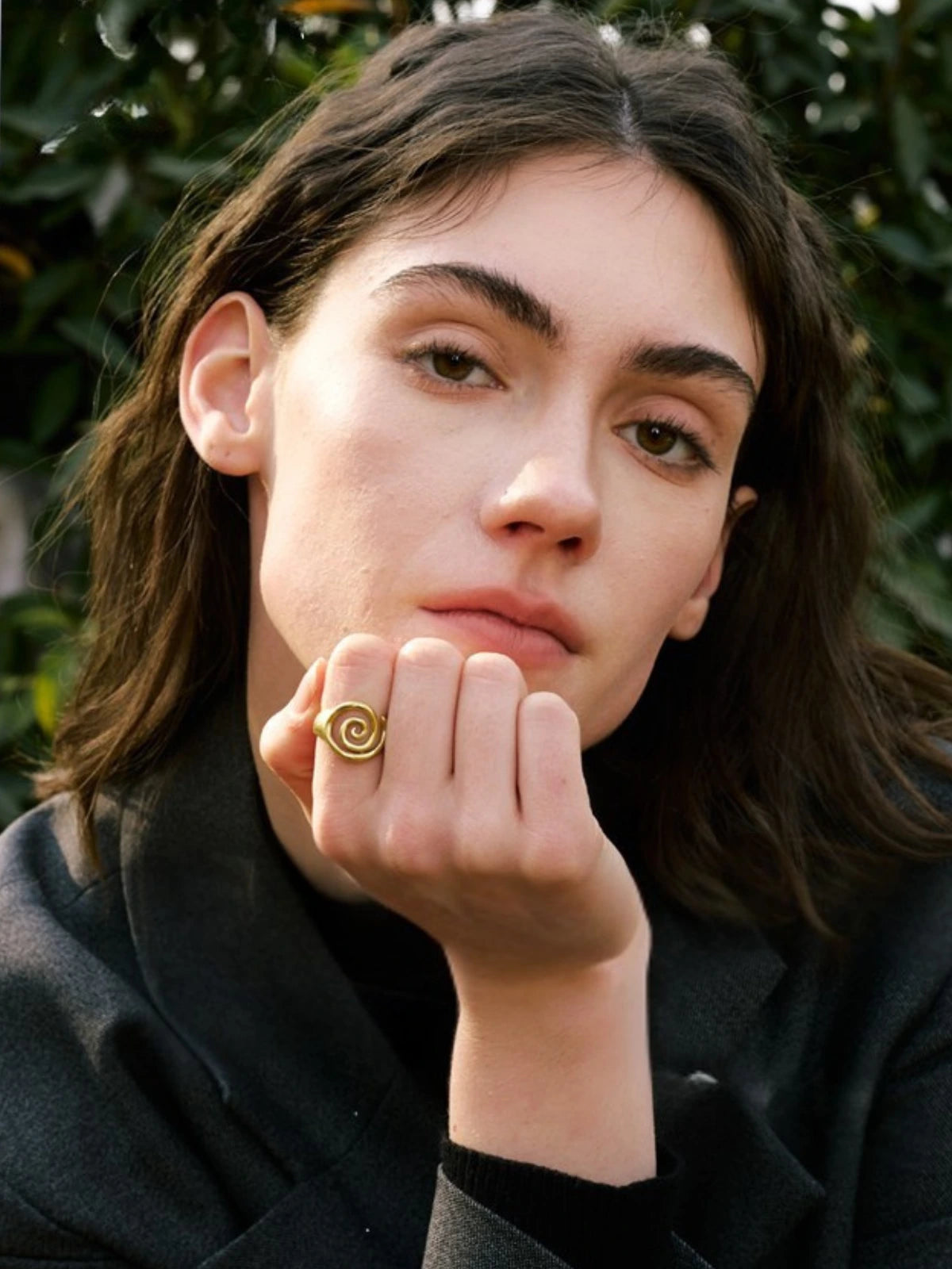 Ophelia Ring worn on finger — 18K gold‑plated stainless steel by Shine With Me, showing elegant design in use.-Woman with a gold ring on her finger, wearing a black coat, against a blurred green background.