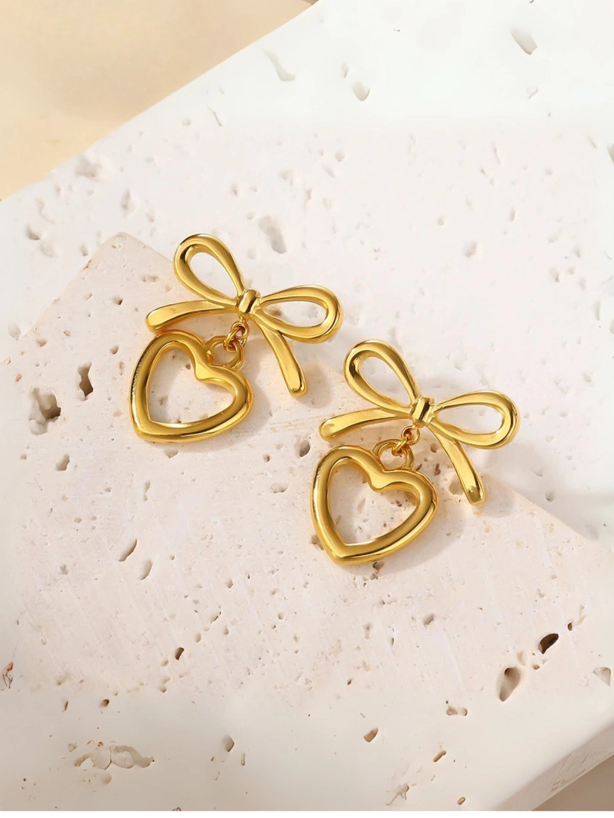 Stefani Bow Earring – Close‑Up Detail-Gold bow and heart-shaped earrings on a textured white surface