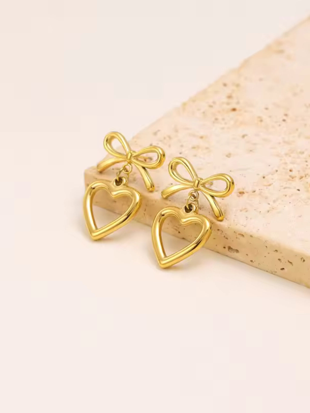 Stefani Bow Earring – Close‑Up Detail-Gold heart-shaped earrings with bow details on a light wooden surface