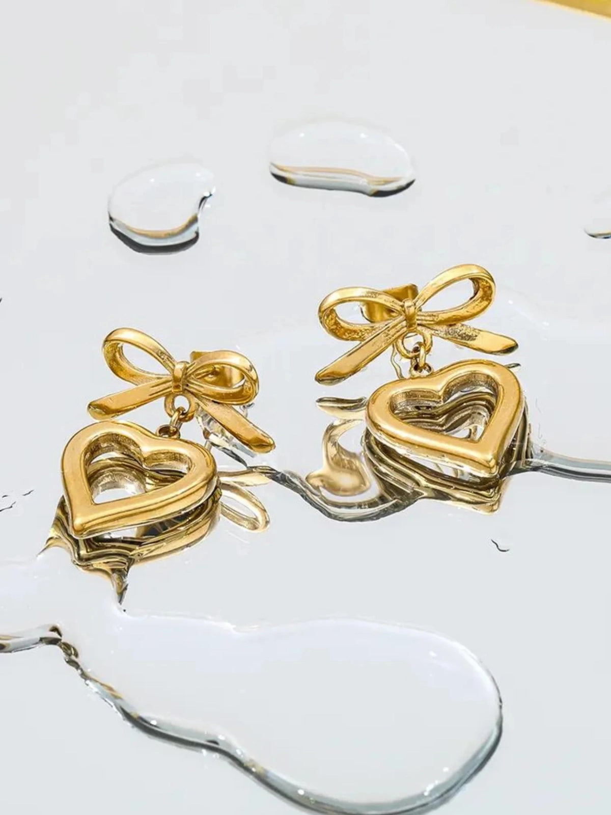Stefani Bow Earring – Close‑Up Detail-Gold heart-shaped earrings with bow details on a clear glass surface
