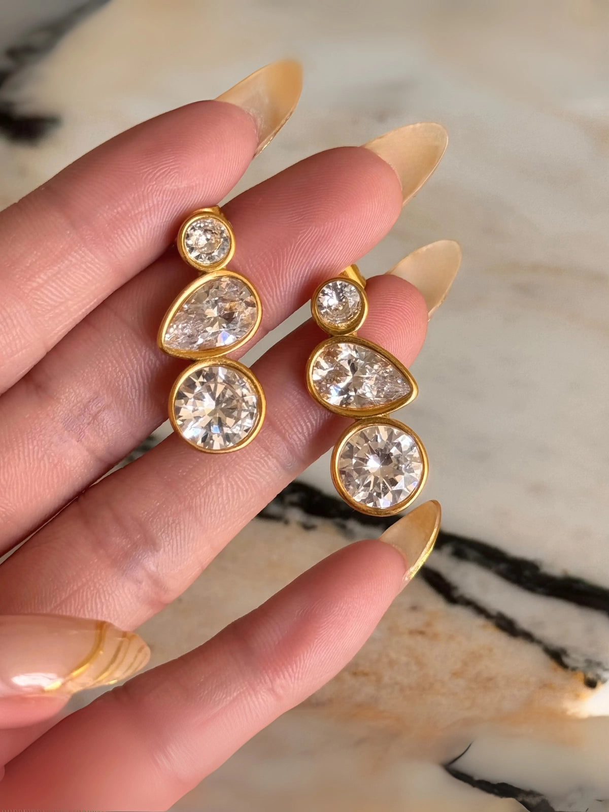 Syndra Earring – Close-Up Detail-Gold and crystal earrings held between fingers against a marble background