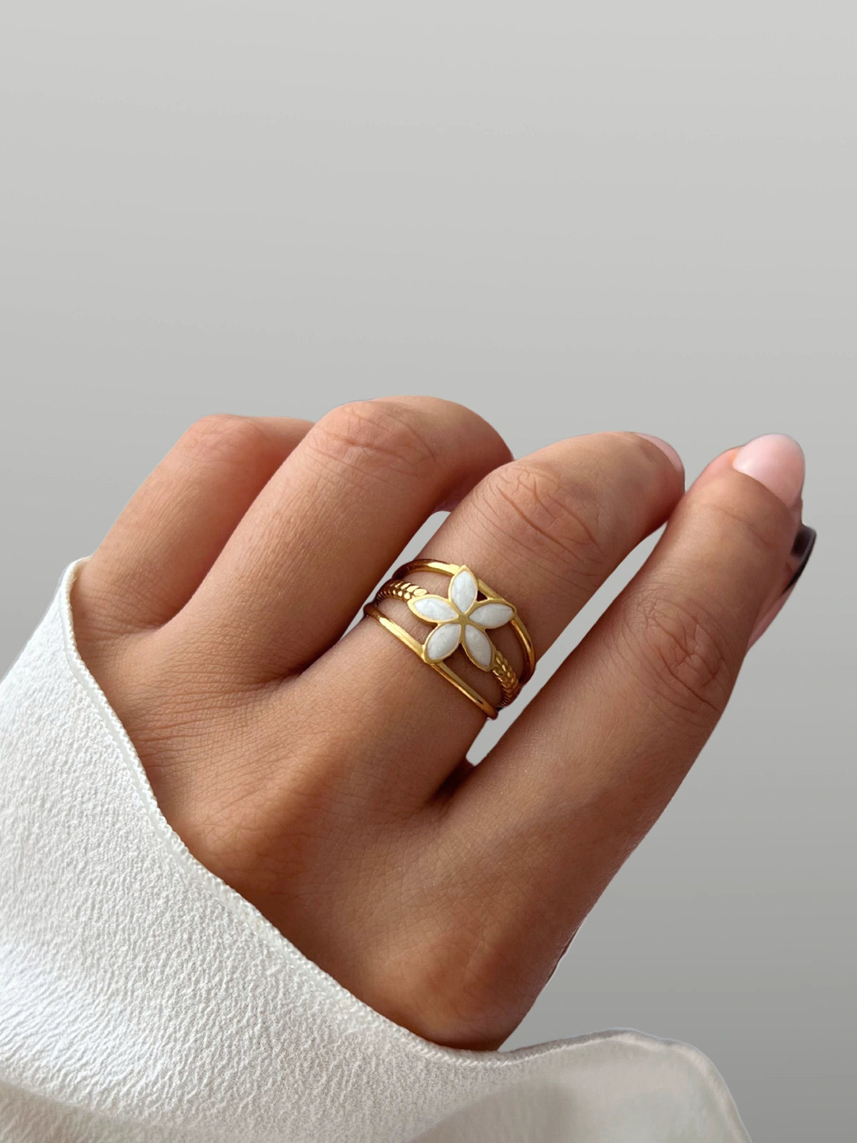 serena-flower-ring-closeup-detail-shine-with-me Hand wearing a gold ring with a white flower design on a neutral background