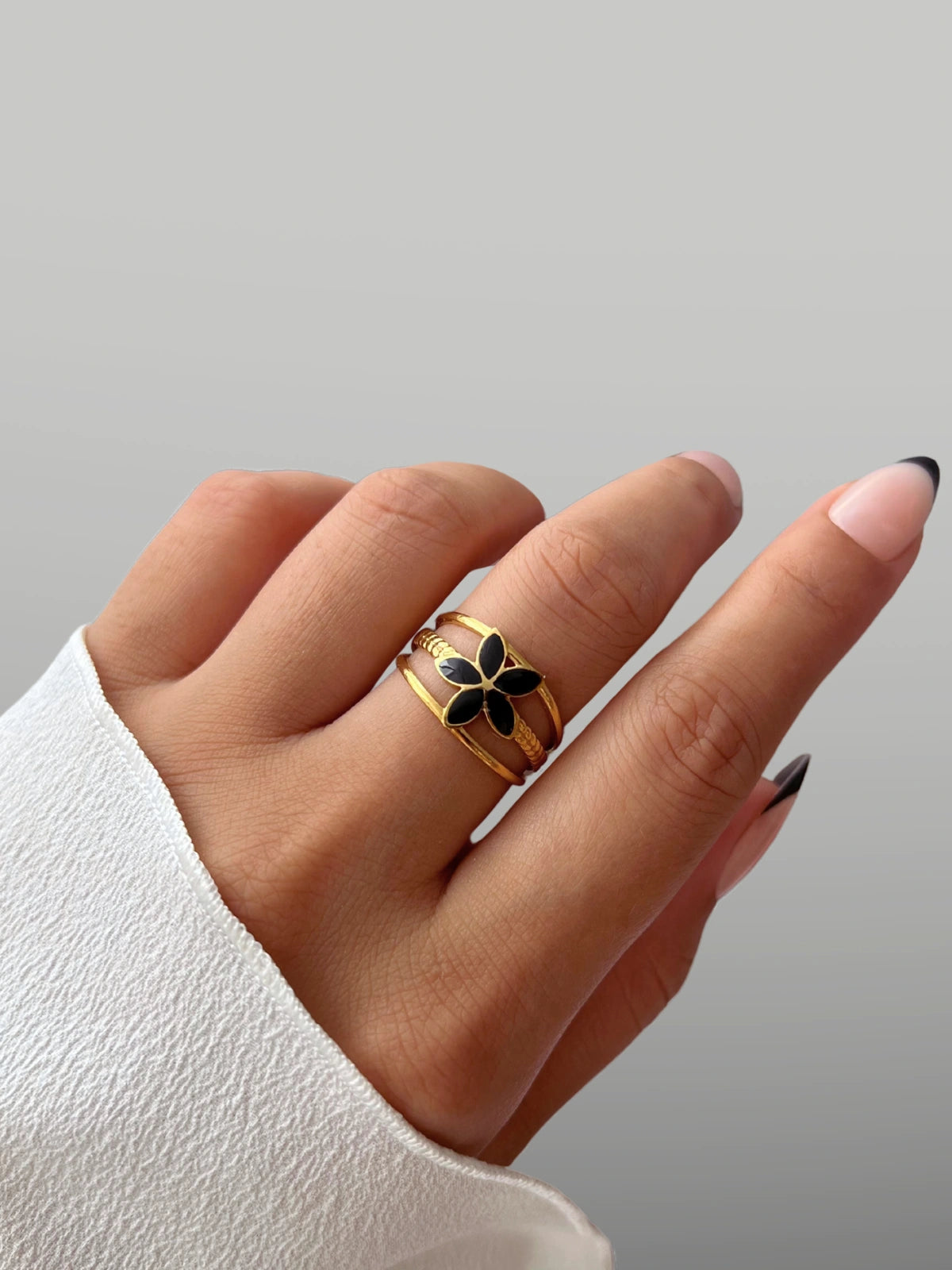 serena-flower-ring-side-angle-shine-with-me Hand wearing a gold ring with a black flower design on a gray background